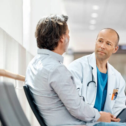 Male doctor counseling mature patient in waiting room. Medical professional is listening to ill man while holding digital tablet in hospital. They are sitting on seat.